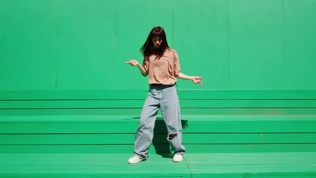 Young woman dancing hip hop on green background wall. Modern dance choreography. Female professional dancer showing dance movings. Musical video.