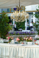 Wedding banquet table with pink rose flowers decoration, crystal chandelier in summer garden, selective soft focus, guests on background. Table set for event, party in outdoor restaurant.
