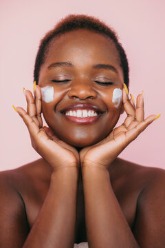 Portrait Of Smiling Black Woman Keeping Her Eyes Closed Applying Face Cream Onto Her Cheeks Isolated Over Pink Background. Beauty Woman Face Skin Care. Facial