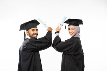 Two graduate students wearing togas holding up certificate while looking back on isolated background