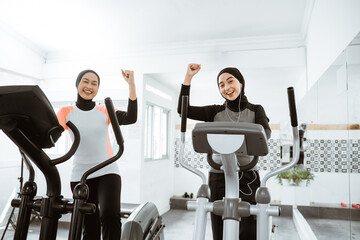 excited beautiful woman with hijab at the gym exercising with friend on static elliptical cycle...