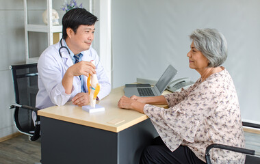 Asian cheerful male professional doctor practitioner in white lab coat with stethoscope sitting smiling holding showing foot skeleton model explaining to old senior female patient in clinic office