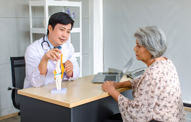Asian cheerful male professional doctor practitioner in white lab coat with stethoscope sitting smiling holding showing foot skeleton model explaining to old senior female patient in clinic office