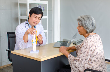 Fototapeta na wymiar Asian cheerful male professional doctor practitioner in white lab coat with stethoscope sitting smiling holding showing foot skeleton model explaining to old senior female patient in clinic office