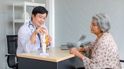 Obraz premium Asian cheerful male professional doctor practitioner in white lab coat with stethoscope sitting smiling holding showing foot skeleton model explaining to old senior female patient in clinic office