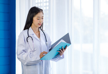 Asian cheerful happy young female professional successful doctor practitioner in white lab coat with stethoscope standing smiling holding reading paperwork document in folder in hospital ward room