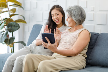 Asian cheerful old senior healthy gray hair female pensioner mother sitting smiling with young daughter on cozy sofa in living room using taplet computer browsing shopping online together