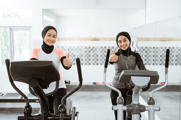 two beautiful woman with hijab at the gym using static elliptical cycle machine and showing thumb up