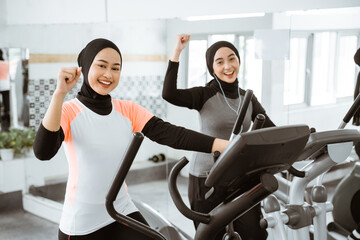 excited beautiful woman with hijab at the gym exercising with friend on static elliptical cycle...