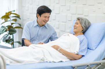 Obraz premium Asian middle aged male son visiting supporting comforting holding hands old senior sick unhealthy pensioner mother patient in blue hospital uniform laying down receiving saline solution drip on bed