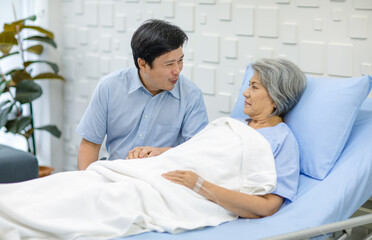 Obraz premium Asian middle aged male son visiting supporting comforting holding hands old senior sick unhealthy pensioner mother patient in blue hospital uniform laying down receiving saline solution drip on bed
