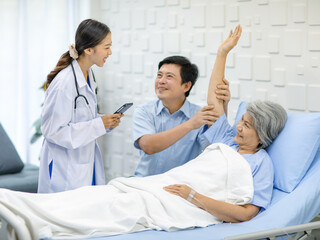 Asian cheerful son holding arm of old senior pensioner mother patient in blue hospital uniform laying down on bed up while professional doctor practitioner in white lab coat visiting in clinical ward © Bangkok Click Studio