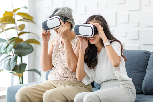 Asian young cheerful daughter wearing VR virtual reality goggles headset sitting smiling on cozy sofa in living room teaching old senior grey hair pensioner mother playing streaming 3d game online