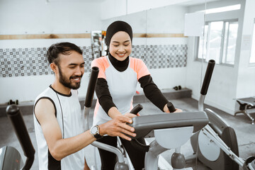 muslim women at the gym doing cardio exercises on static elliptical cycle machine with friend help...