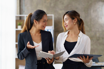 Two beautiful Asian business women stand and talk about work using a tablet in the office.