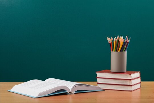 Stack of books and holder with stationery on table near chalkboard in classroom