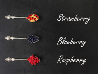 Three spoons with berries (raspberries, blueberries, strawberries) on a black background with inscriptions. Close-up. Flatlay.