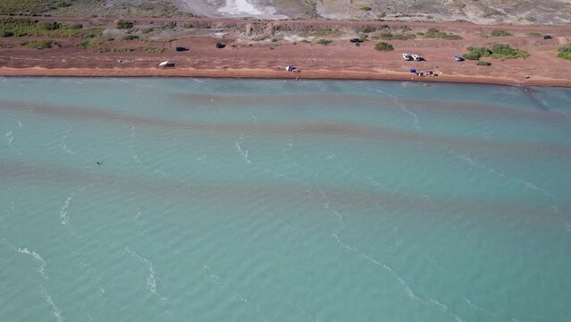 A beach with blue water color and sandy waves. Top view from a drone of vacationing people, tents and cars. Brown sand of Lake Balkhash. Blue sky. Bushes and several trees grow. Family vacation by sea