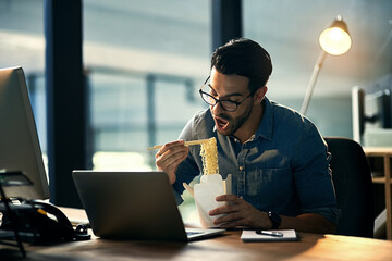 Young businessman working late and eating at desk. Man having takeout food in the office at work station in the evening. Male worker feeding his brain from work during the night at the workplace.
