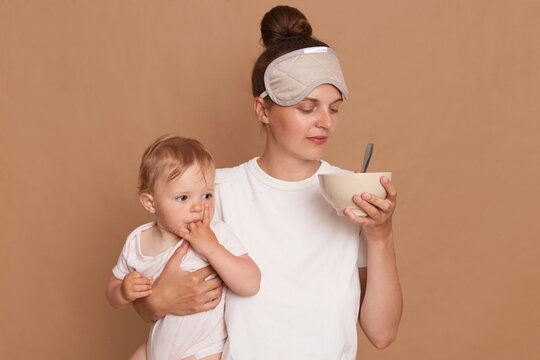 Portrait Of Woman Wearing White T Shirt And Sleeping Mask, Holding Infant Kid In Hands, Smelling Food In Plate For Feeding Baby, Posing Isolated Over Brown Background.