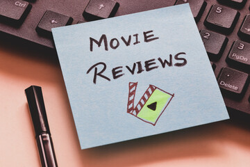 Sticky note with the handwritten text Movie Reviews and film movie icon on keyboard. Close-up image.