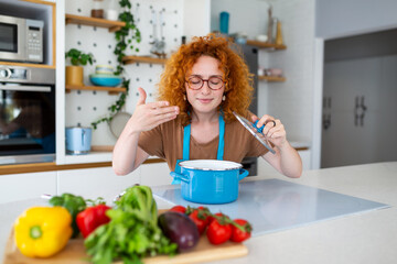 Cheerful young pretty female in apron prepare lunch and smell dish in kitchen at home. Woman cooking dinner for family at new recipe at home.