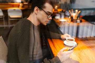Young man smiling while drinking coffee and using cellphone in cafe