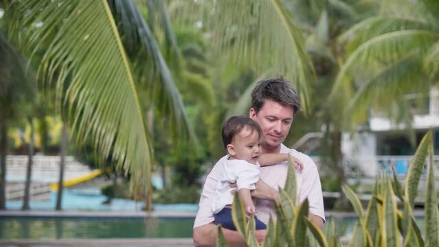 Not far from the pool, a young dark-haired handsome father in summer clothes approaches green beautiful flowers with his cute child in his arms. Holidays with children on vacation.