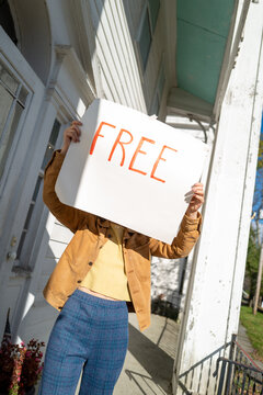 A person in vintage clothes holds up a handmade sign that says 'free' on a porch 