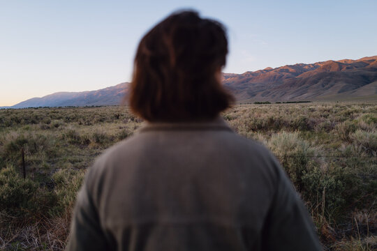 A young person with red brownish hair in a brown jacket gazes longingly at dry mountain landscape