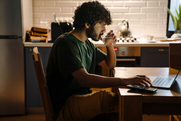Young curly indian man drinking water while working with laptop