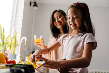 Young smiling asian mother looking on her little daughter cooking