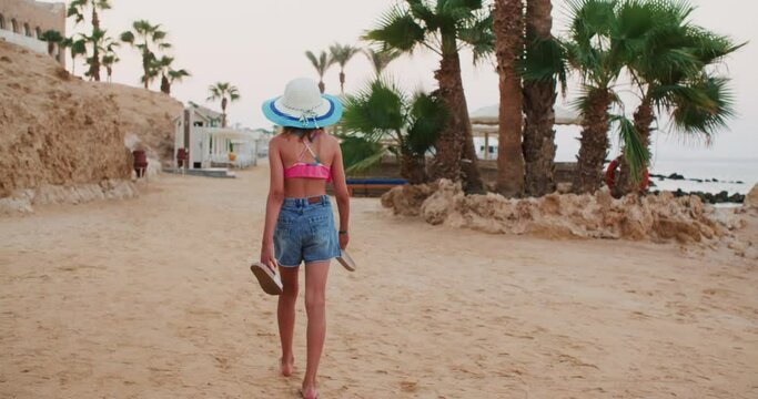 Traveling, Tourism, Resting on Vacations Concept. Teen Girl is Leaving the Beach in Tropics Carrying Slates in her Hands. Seascape with Sand, palm trees and Rocks. 
