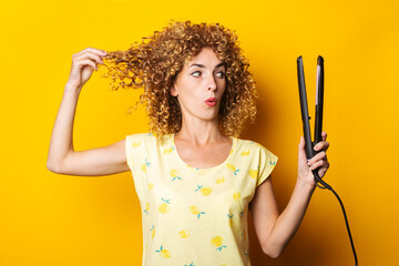 young woman with curly hair looks in surprise at the hair straightener on a yellow background
