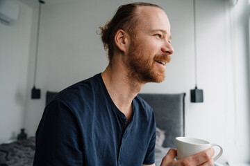 Young ginger man drinking coffee while sitting on bed