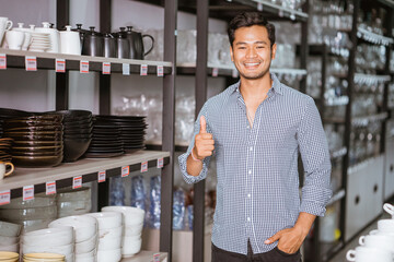 Handsome asian man standing with thumbs up beside shelf in homeware store