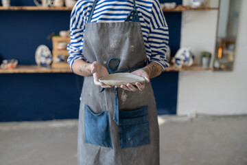 Cropped photo of woman ceramic artist in dirty apron holding handmade pottery item in hands, potter...