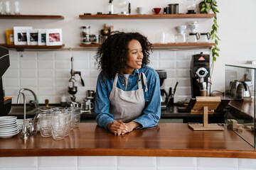 Black young barista woman wearing apron smiling while working in cafe