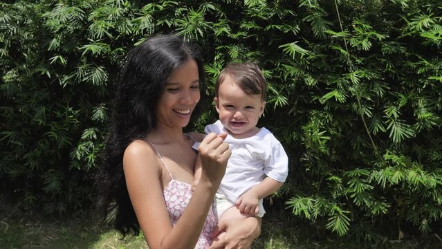 A dark-haired woman stands in a green garden with a small child in her arms. Child from 8-9-10 months. Woman clenching her hand into a fist smiling. The child watches the movement of the mother.