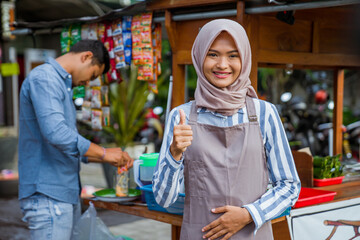 proud young muslim woman standing in front of her small traditional food shop and showing thumb up...
