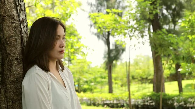 Heartbroken asian woman unhappy sadness in love spends her time alone relax in the garden contemplative,leaning against tree, looking at something around you and  calmness giving yourself time.
