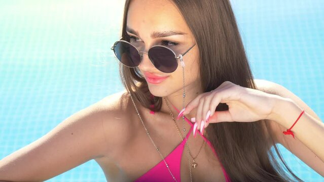 Stunning young woman wearing sunglasses in a blue outdoor swimming pool, luxury vacation, leisure at the beach concept, summertime patadise in a swimming pool