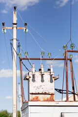 A metal rusty white elecrical transformer with wires going towards it agaisnt a blue clear sky and an electrical pole