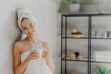 Pretty smiling woman with healthy skin wrapped in white towel, stands glad indoor, drinks coffee,...