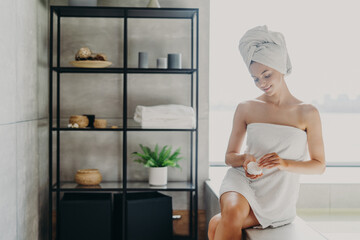 Daily routine, wellness and hygiene concept. Pleased young slim young European woman wrapped in bath towel applies body cream, has healthy skin, poses in cozy bathroom, takes care of herself.