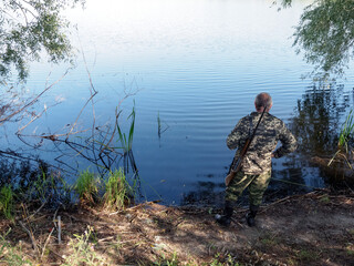 A hunter in camouflage clothes with a rifle and rubber boots stands near the lake and looks into...