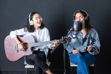 Naklejka na ściany i meble Happy cheerful pretty smiling of portrait Two young Asian woman vocalist Wearing Headphones with a guitar recording a song front of microphone in a professional studio
