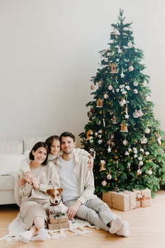 Three family members pose on floor in cozy room, gift boxes around, decorated New Year tree and sofa. Father, mother and daughter with jack russell terrier dog anticipate for coming holiday.