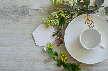 Flatlay with an empty cup on a saucer with mockup an empty white card framed by branches of green leaves, white lilac flowers and yellow small flowers