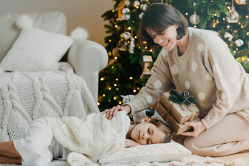Obraz na płótnie Canvas Famaily, happy winter holidays concept. Caring mom stands on knees near sleeping daughter, prepares presents for New Year, has pleasant smile, pose in cozy living room. Christmas night and miracle